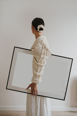 Woman holding large frame mockup with blank poster on minimalist neutral wall background