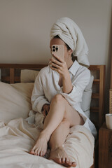 Lifestyle Portrait Of Woman Sitting On Bed Using Mobile Phone With Head Wrapped In Towel