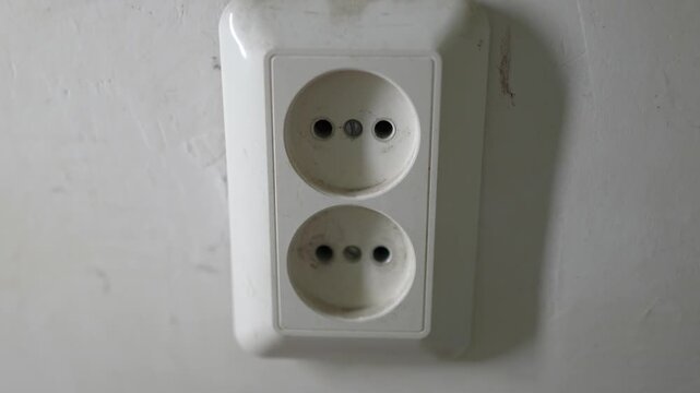 Close-up shot of a white European wall power outlet on a light-colored wall. A human hand plugs a white power adapter into the lower socket. 