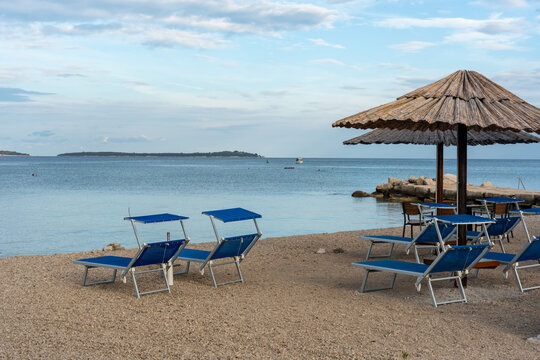 Blue beach sunbeds with canopy shades and rustic thatched umbrella on quiet pebble adriatic shore with calm sea, rocks and distant island on cloudy summer morning. Concept of relaxing coastal holiday.