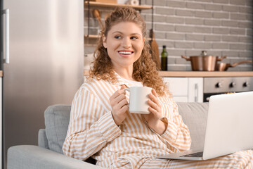 Pretty young woman with white cup of coffee and laptop sitting on sofa in kitchen