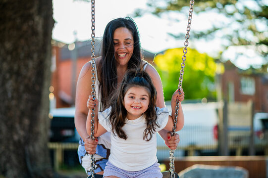 A woman pushes her daughter on a swing at a park during a sunny day in the afternoon.