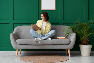 Pretty young woman reading book on sofa near green wall
