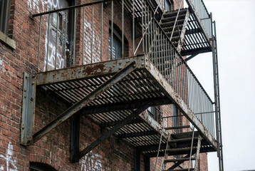 Rusty metal fire escape and industrial balcony attached to an old red brick building. Urban decay and exterior city architecture with emergency stairs and graffiti.