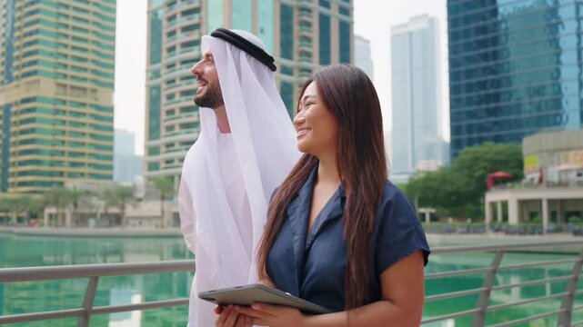 Arab businessman in traditional kandura and Asian real estate agent woman, a professional holding a tablet, walking in a modern Dubai city.