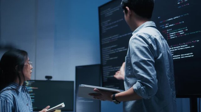 Asian Male developer leading a team meeting, pointing to code on a big monitor while reviewing an AI chatbot module. Concept of collaboration, debugging for coding teamwork in a tech company office.