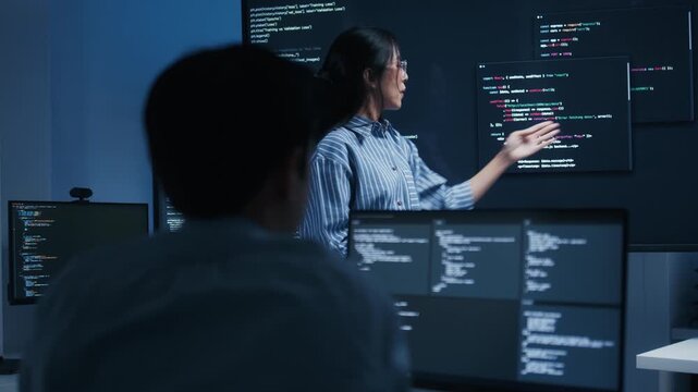 Asian Female Developer Leader pointing to monitor, presenting code on screen, explaining code to colleagues Male Developer coworker asking and discussing working together collaboration in office.