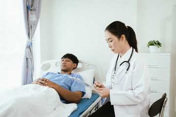 Fototapeta premium Female doctor reviewing medical records on a clipboard while a male patient rests in a hospital bed during inpatient care.