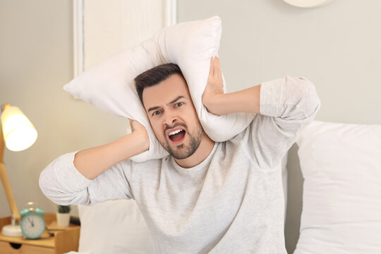 Young man with pillow suffering from loud neighbours in bedroom