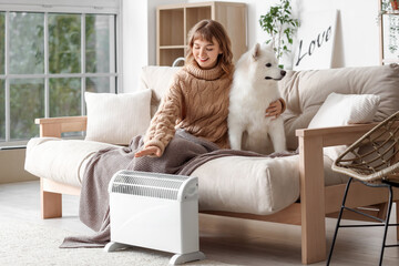 Young woman with blanket and Samoyed dog on sofa warming near radiator at home