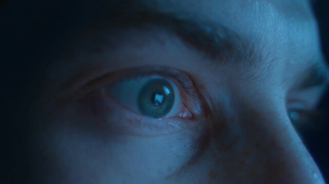 Man eyes with a crazy look nervously staring at the screen with a reflection of social media news feed at night. Harm of excessive content viewing, anxiety, digital addiction, doomscrolling.