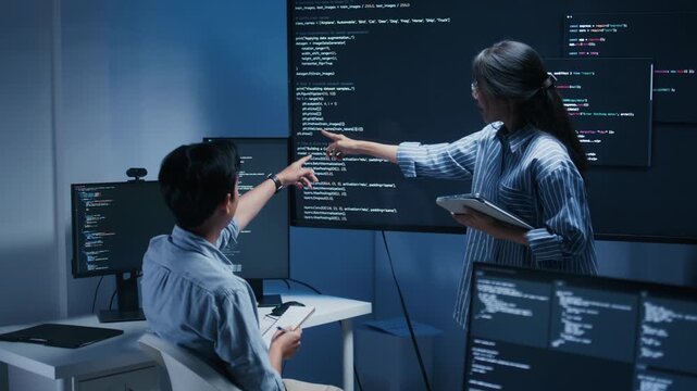 Professional Female Computer Programmer explaining code on monitor while male software developer listening, discussing and brainstorming working together during software development meeting in office.
