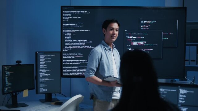 Asian Male Software Engineer presenting code on Screen while Holding Digital tablet and Explaining code, Ai Chatbot Feature, Software Development Task to Colleagues while Collaboration in tech office.