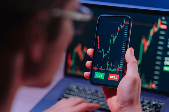 Over shoulder view of crypto trader analyzing live crypto and stock charts on smartphone with trading platform app, displayed on laptop and phone screen in dark neon lit room.