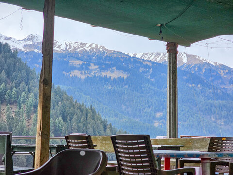 Horizontal Landscape View of Outdoor Cafe Balcony with Plastic Chairs Overlooking Dense Pine Forest and Snowy Himalayan Peaks in Himachal Pradesh India.