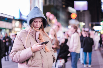 Fototapeta premium Teenage girl using smartphone at Times Square New York at night, young woman in winter hooded jacket standing among crowd and neon lights in busy urban street