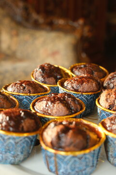 Close-up shot of freshly baked moist chocolate muffins in blue and gold patterned cups with chocolate glaze, featuring warm sunlight and a beautifully blurred vintage background.