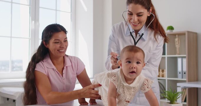 Smiling mother holds her baby at pediatric checkup as the pediatrician listens with a stethoscope in medical clinic, focusing on health and care. Family doctor does examination with a stethoscope.