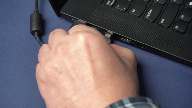 Hand connects USB-C cable to laptop port while other cables and USB devices are arranged on a blue surface in a well-lit workspace