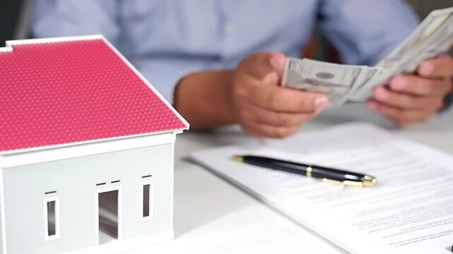A man counts dollar bills with a miniature house in front of him. The concept of renting a house or paying taxes on an apartment. The first payment on a mortgage, rent, and home insurance.