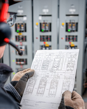 A technician wearing gloves and safety glasses studies an electrical schematic in front of a control panel with illuminated indicators.