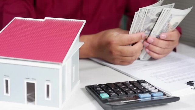 A man counts dollar bills with a miniature house in front of him. The concept of renting a house or paying taxes on an apartment. The first payment on a mortgage, rent, and home insurance.
