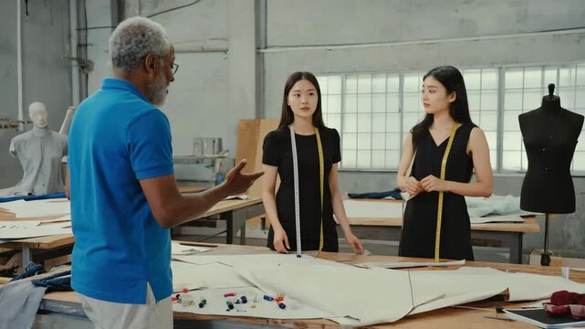Senior african male fashion designer mentoring two young asian female students in a workshop. Professional tailor teaching apprentices about pattern making and clothing creation