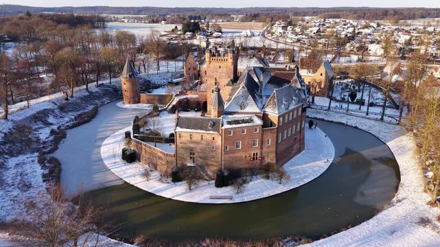Aerial golden hour view of a historic moated castle in winter. Snow dusts the brick fortress and surrounding village, while long shadows stretch across the partially frozen circular moat.