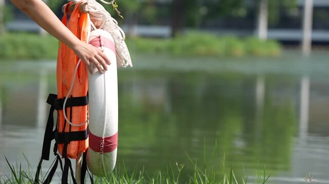 Lifebuoy and life jacket rescue safety gear at water lake shoreline, rope emergency flotation ring held near grass, lifeguard readiness for outdoor summer freshwater response