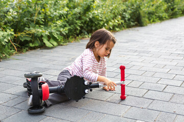 Little girl sitting on the pavement after a fall, holding her scooter with curiosity and calmness, surrounded by greenery, representing childhood, learning, and resilience through play