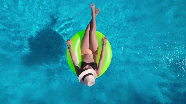 Aerial view of young woman in hat relaxing in swimming pool, beautiful girl in bikini floats on inflatable ring donut and has fun in water, tropical vacation on holiday resort, top drone view