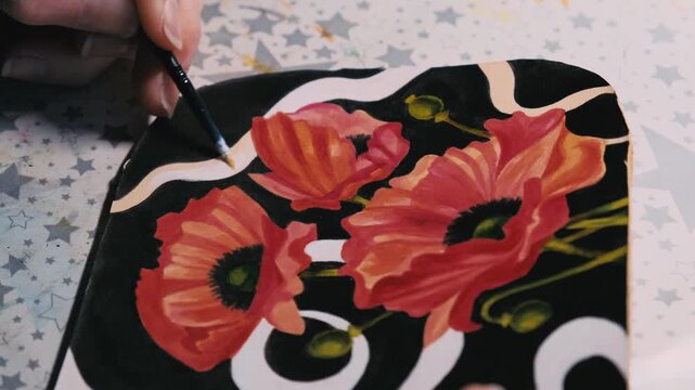 Close-up of a hand painting a red poppy flower on natural vegetable-tanned leather. Detailed brushwork on genuine cowhide surface, showing texture, color layering and craftsmanship.