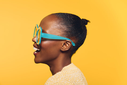Profile of a smiling African woman with dark skin and hair in a bun wearing large blue sunglasses and a light sweater. Bright yellow background creates a cheerful and modern mood.