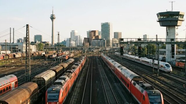 A bustling train yard with multiple tracks and red and white trains in the foreground City skyline with a tower and modern buildings in the background under a bright sky