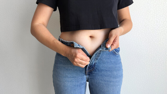 Young woman in black crop top trying to button tight blue jeans, close up view of slim waist and bare abdomen against neutral background, weight loss and body shape control concept