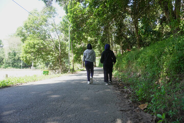Fototapeta premium Two young Muslim women walking together for morning exercise along a green park pathway filled with trees on a bright sunny day. 