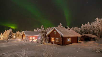 Experience the magic of winter in Akaslompolo, Lapland, where colorful northern lights illuminate rustic cabins nestled in snow-covered landscapes. This serene scene captures holiday enchantment. © Fokke Baarssen