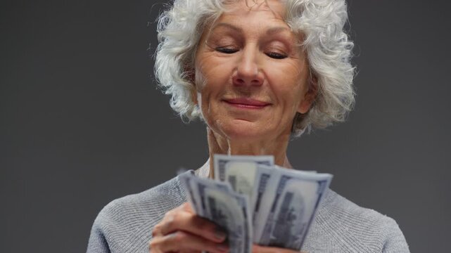 Portrait: Happy senior woman with grey hair counting USD banknotes. Rich old lady getting rich with jackpot. Face of mature female person holding cash money paper dollars. Wealth retirement concept