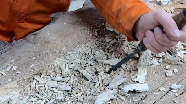 A carpenter in overalls chiselling a wooden slab in a woodworking shop. A person working. Tools, chisel, mallet. Woodworking. Sawdust, wood chips. Real people. Lifestyle, Authentic, Candid