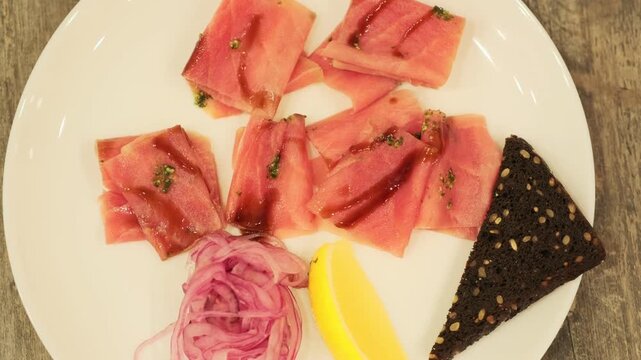 Tuna stroganina with pickled onion, lemon wedge, and rye bread on a white plate. Fresh seafood for gourmet dining, top view.