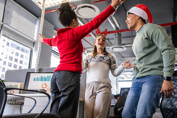 Diverse coworkers celebrating near monitors showing charts in office wearing Santa hat and antlers