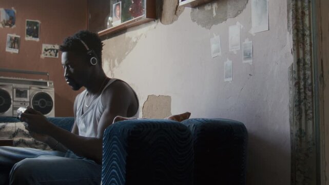 Young Black man sitting on sofa, wearing headphones and listening to music from retro cassette player, relaxing in modest living room with vintage audio equipment and personal photos on peeling walls