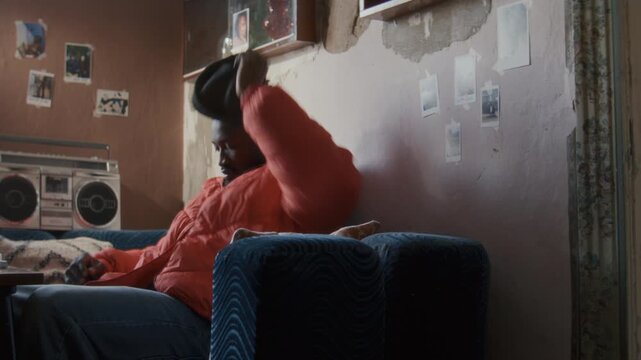 Young African American man walking in modest worn apartment with peeling walls and vintage stereo, sitting on couch, counting cash and reflecting on financial situation