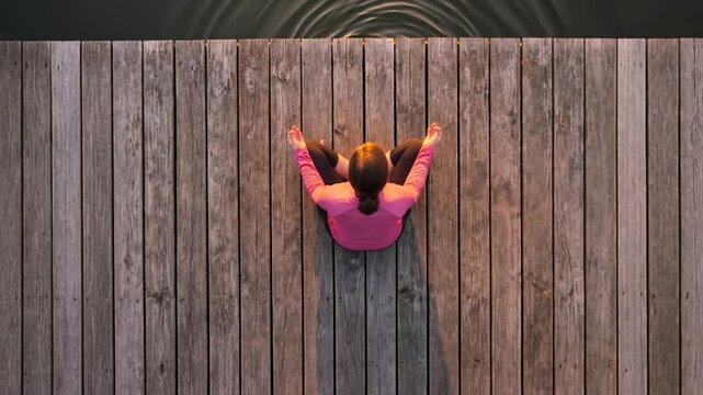 Aerial top-down view of a woman meditating on a wooden dock surrounded by water