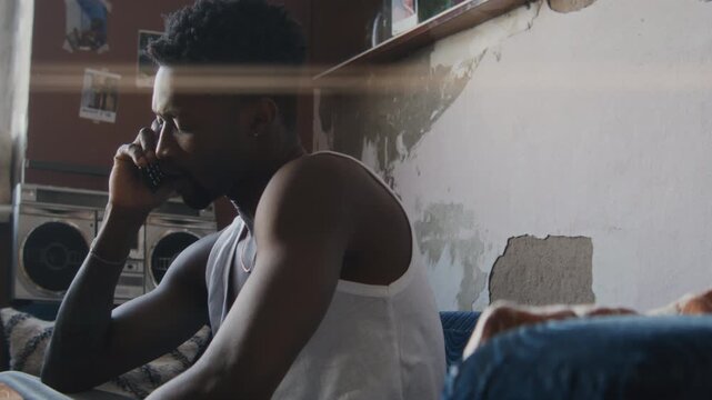 Young Black man sitting on sofa in modest living room with worn textured walls, dialing number on flip phone and talking seriously, bright sunlight streaming through window