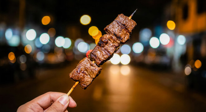 Hand Holding Grilled Anticucho Skewer With City Night Bokeh