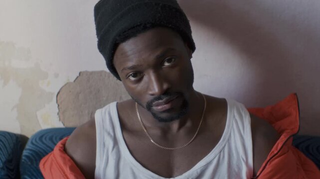 Young serious Black man wearing beanie and streetstyle outfit sitting on couch against worn plaster wall, looking at camera and moving his head to music beat. Video portrait
