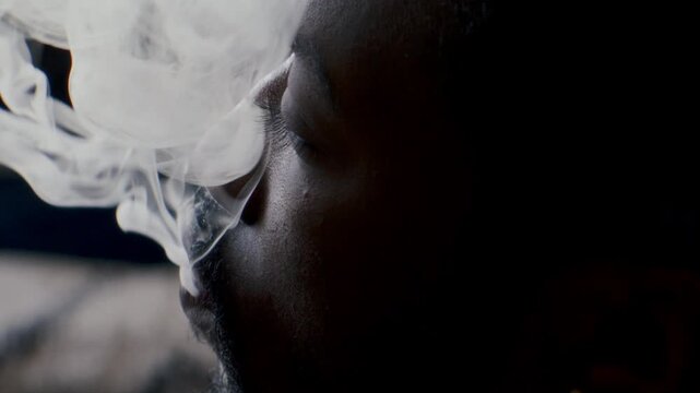 Close up view of young Black man smoking joint, exhaling dense cigarette smoke and looking away pensively