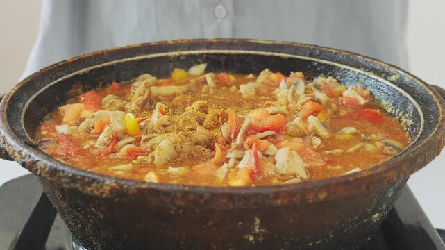 In a deep cast-iron skillet, vegetables and chicken are simmering in sauce and spices, stirred with a spoon. Medium shot.