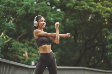 Smiling Asian woman athlete wearing headphones in sports bra stretching her arms before a workout in a green park. Healthy lifestyle fitness and outdoor exercise concept.